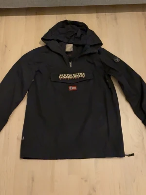 Svart vindjacka från Napapijri - Stilren svart vindjacka från Napapijri med huva och dragkedja vid halsen. Jackan har en stor ficka framtill med logotyp och norsk flagga, samt patch på ärmen. Perfekt för blåsiga dagar och enkel att matcha.