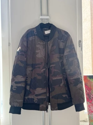 Camo bomberjacka från Calvin Klein - Snygg bomberjacka från Calvin Klein i camouflage-mönster. Jackan har svarta muddar vid ärmslut och midja, dragkedja framtill och en klassisk bomberkrage. Quiltad design och ficka med dragkedja på ärmen. Perfekt för dig som vill sticka ut.