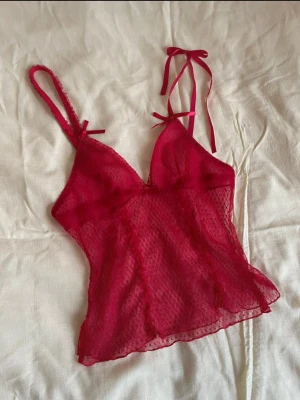 Röd mesh linnetopp med rosetter - Söt röd mesh topp med tunna axelband och små rosetter framtill. Toppen är lätt transparent med diskret prickigt mönster och har en feminin, figurnära passform. Perfekt för att addera en romantisk vibe till din outfit. Superfin festtopp