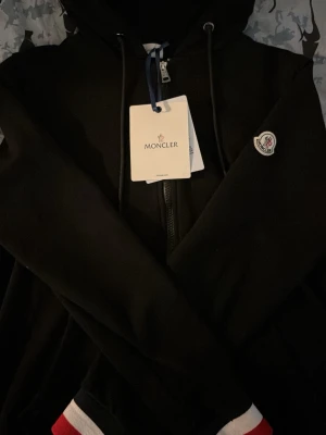 Svart Moncler hoodie med dragkedja - Svart hoodie från Moncler med dragkedja framtill, huva med snören och Moncler-logga på bröstet. Ärmsluten har snygga ränder i rött, vitt och blått. Perfekt för dig som vill ha en stilren och exklusiv look.