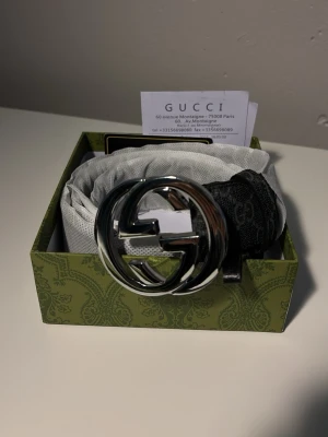 Gucci svart bälte med silverdetalj - Snyggt svart bälte från Gucci med stor, rund silverspänne i form av GG-loggan. Bältet har diskret GG-mönster längs hela bandet och är tillverkat i skinn. Perfekt accessoar för att lyfta din outfit.