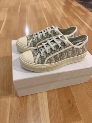 Dior skor walk’n dior  - Bild 2 kvitto. Säljer ett par walk’n dior låga sneakers i grått och vitt med klassiskt Dior-monogram på canvas. Skorna har vita snören, gummisula med stjärndetalj undertill och tåhätta i gummi. Perfekta för dig som vill ha en clean och lyxig look.   
