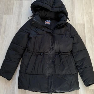 Svart pufferjacka med huva - Snygg svart pufferjacka från Jacket of Design med stor huva och dragsko i midjan. Jackan har två stora fickor framtill och stängs med både dragkedja och knappar. Perfekt för kalla dagar och ger en riktigt cool streetwear-look.