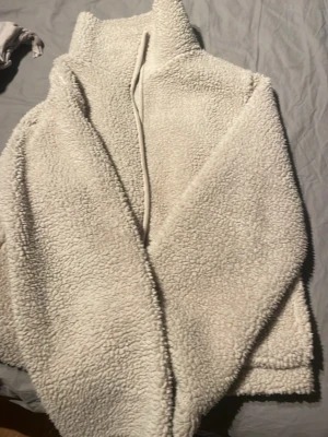 Vit teddyjacka med hög krage - Mysig vit teddyjacka i fluffigt material med hög krage och dragkedja framtill. Jackan har en avslappnad passform och är perfekt för kalla dagar. Materialet är mjukt och ger en trendig look.