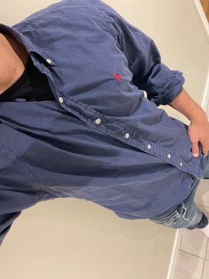 Blå skjorta från Ralph Lauren - Klassisk blå skjorta från Ralph Lauren med knappslå och vit knappstängning. Skjortan har lång ärm och den ikoniska röda logotypen broderad på bröstet. Tillverkad i bomull och har en normal passform. Perfekt till jeans eller chinos.💋Modellen bär storlek L och är 181 cm lång och väger 76 kg kom gärna vid frågor eller funderingar💋har en även i storlek S