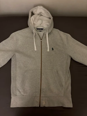 Grå zip-hoodie från Polo Ralph Lauren - Snygg grå hoodie med dragkedja från Polo Ralph Lauren. Klassisk design med huva och vita snören, två fickor framtill och broderad logga på bröstet. Mjuk bomullsblandning som är perfekt för en chill och avslappnad stil.