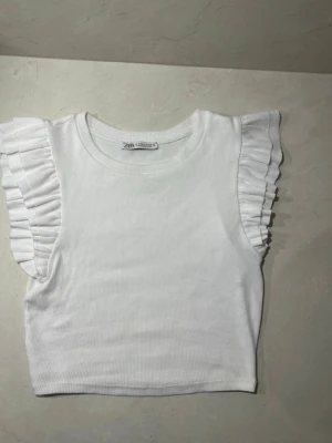 Vit ribbad topp med volangärmar Zara - Superfin vit ribbad topp från Zara med volangdetaljer på axlarna. Ärmlös modell med rund halsringning och croppad passform. Perfekt att styla med jeans eller kjol för en trendig look.