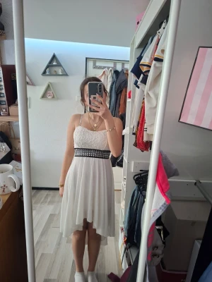 Vit spetsklänning med asymmetrisk kjol - Säljer en jätte fin vit klänning med spetsliv utan axelband. Kjolen är asymmetrisk och faller lätt, midjan markeras av ett svart band med nitar. Baksidan har spetsdetaljer och klänningen är ärmlös. Perfekt för dig som gillar en romantisk och trendig look. Står ingen storlek men skulle typ säga storlek S. Aldrig använd.