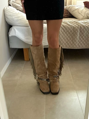 Beiga cowboy boots med fransar - Säljer ett par höga cowboy boots i beige mocka med långa fransar längs en av sidorna och dekorativa avtagbara spännen med metall-detaljer runt ankeln. Bootsens tå är lätt fyrkantig och sulan är svart. Perfekta för dig som vill sticka ut med westernstil. Det står att storleken är 37 men de är stora och passar med med 37,5-38! 