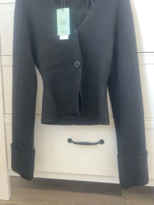 Svart kavaj liknande tröja  - Stilren svart kavaj liknande med två knappar framtill och breda, uppvikta ärmslut. Kavajen har en något croppad passform och markerad midja. Materialet är mjukt och lite tjockare, perfekt för en trendig look. Inte använd bara provad. Prislappen är kvar.