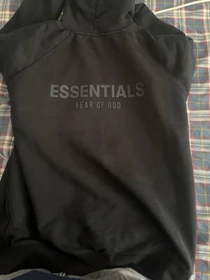 Svart Essentials Fear of God hoodie - Säljer en svart hoodie från Essentials Fear of God med stor logga på ryggen och mindre logga på bröstet samt ärmen. Hoodien har huva och en stor magficka. Materialet är mjukt och bekvämt, perfekt för en avslappnad look. Pris kan diskuteras vid snabb affär!