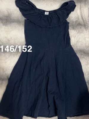 Marinblå klänning med volang - Snygg marinblå klänning med volangdetalj upptill och offshoulder-design. Klänningen har en svängande kjol och är tillverkad i ett lätt material som passar perfekt till varmare dagar.🌸 Fint använt skick. Storlek 146/152