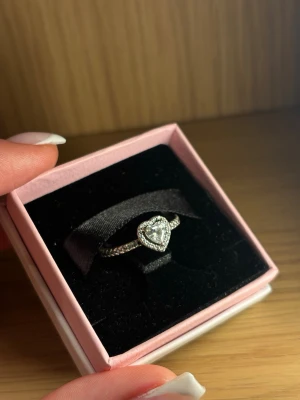 Pandora silverring med hjärta - Säljer en snygg silverring från Pandora med ett glittrande hjärta i mitten. Ringen har små stenar längs bandet och hjärtat är inramat av gnistrande detaljer. Perfekt för dig som gillar romantisk och elegant stil.