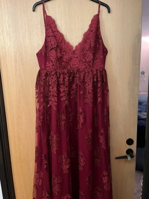 Vinröd långklänning med spetsdetaljer - Balkänning. Elegant vinröd långklänning med tunna axelband och v-ringning. Klänningen har vackra spetsdetaljer och blommigt mönster över hela tyget. Perfekt för speciella tillfällen och har en romantisk känsla.