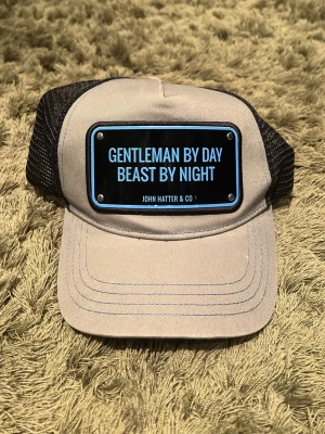 Grå och svart keps John Hatter & Co - Cool grå keps med svart mesh och justerbar snapback från John Hatter & Co. Framtill sitter en metallskylt med texten 'Gentleman by day, beast by night'. Perfekt för dig som vill sticka ut med en unik accessoar.