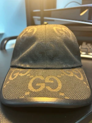 Svart stilren keps med GG-mönster - Svart keps med genomgående GG-mönster över hela tyget som ger ett stilrent och exklusivt utseende. Modellen har böjd skärm med matchande mönster framtill och en mjuk, strukturerad passform. Justerbart band baktill med spänne för enkel anpassning av storleken. Enkel att matcha med olika outfits tack vare den helsvarta designen och de diskreta detaljerna.