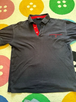 Mörkblå piké från Polo Ralph Lauren - Snygg mörkblå pikétröja från Polo Ralph Lauren med små diskreta prickar, röd knappslå och röd broderad logga på bröstfickan. Pikén har korta ärmar och kontrastdetaljer i rött vid krage och ficka. Perfekt för en stilren och avslappnad look.