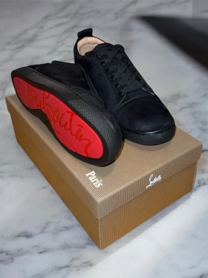 Svarta Louboutin Sneakers - 43 - Snygga svarta sneakers från Christian Louboutin i mocka med klassisk röd sula och svarta skosnören. Låg modell med diskret logga och stilren design som sticker ut med den ikoniska röda undersidan. Skorna kommer med paket, certifikat och även den originella dustbag.