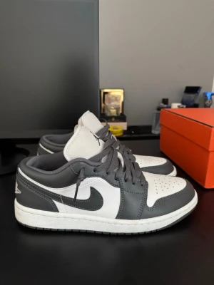 Air Jordan 1 Low grå och vita sneakers - Snygga Air Jordan 1 Low sneakers i grått och vitt läder med klassisk swoosh på sidan och perforerad tå. Skorna har grå snörning, vit mellansula och diskret Jordan-logga på plösen. Perfekta för dig som gillar streetwear och vill sticka ut med stilrena färger.