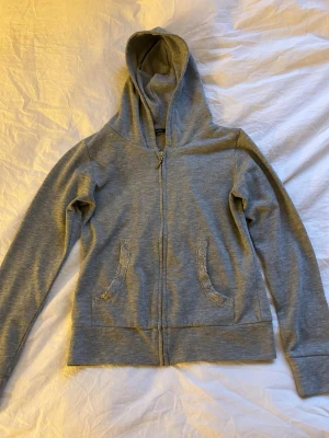 Ljusgrå hoodie med spetsdetaljer - Ljusgrå hoodie med dragkedja och huva. Fickorna har snygga spetsdetaljer som ger en extra touch. Mjuk bomullsblandning och ribbade muddar vid ärmslut och nederkant.