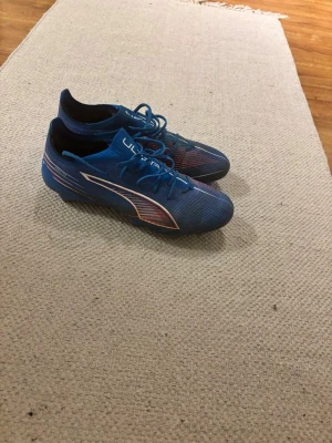 Blåa Puma fotbollsskor med dobbar - Säljer ett par blåa fotbollsskor från Puma med vita och röda detaljer. Skorna har snörning och är tillverkade i syntetmaterial med låg profil och fasta dobbar för spel på gräs. Perfekta för dig som vill ha bra grepp och stil på planen.