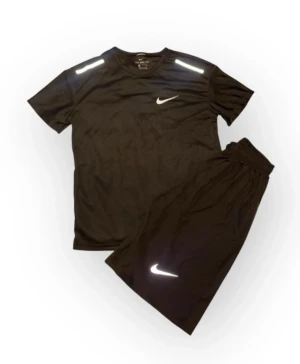 Svart Nike träningsset med reflex - Säljer ett svart träningsset från Nike med t-shirt och shorts. Båda plaggen har vita Nike-loggor och reflexdetaljer på axlar och ben. Materialet är lätt och andas, perfekt för sport och träning. Elastisk midja på shortsen för skön passform.