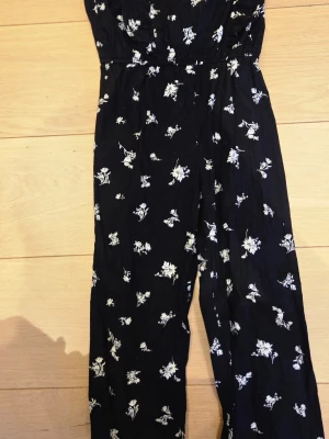 Svart blommig jumpsuit Vintage Stories - Snygg svart jumpsuit från Vintage Stories med vitt blommönster. Jumpsuiten har långa ben och markerad midja, perfekt för en trendig look. Tunt och lätt material som känns skönt mot huden. Passar dig som gillar en avslappnad men stilren stil.