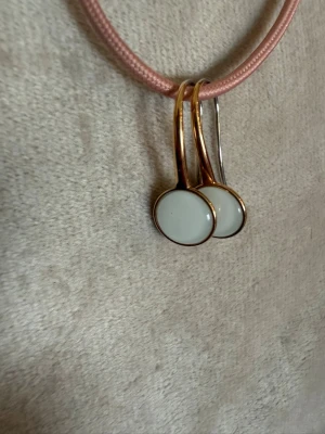 Guldiga örhängen med vit rund detalj - Snygga örhängen med rund vit detalj infattad i guldfärgad metall. De har en enkel och stilren design med krok som gör dem lätta att bära. Perfekta för dig som gillar minimalistiska smycken med en touch av elegans.