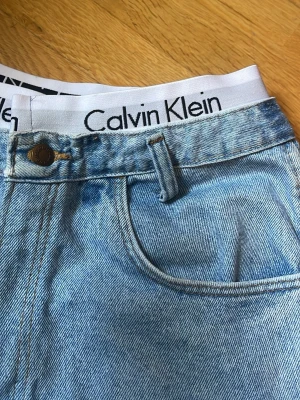 Wrangler jeansshorts med Calvin Klein-detalj - Säljer ett par ljusblå jeansshorts från Wrangler med råa fransade kanter och klassiska bakfickor. Shortsen har hög midja och en synlig Calvin Klein-resår i midjan för en unik look. Perfekta för sommaren! Medium size 