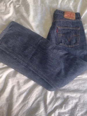 Levi's 501 blå raka jeans - Klassiska blå Levi's 501 jeans med rak passform och fem fickor. Jeansen har den ikoniska röda Levi's-lappen på bakfickan och patch i läder bak i midjan. Tillverkade i slitstark denim, perfekta för en avslappnad och tidlös stil.