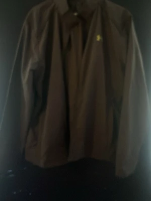 Svart vindjacka från Under Armour - Snygg svart vindjacka från Under Armour med klassisk logga i gult på bröstet. Jackan har dragkedja framtill och är tillverkad i ett lätt och smidigt material som skyddar mot vind. Perfekt för aktiva dagar eller när du vill ha en sportig look.