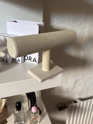 Beige smyckesställ i sammet - Stilrent smyckesställ i beige sammet med T-formad design. Perfekt för att organisera armband, klockor eller halsband. Stället har en stadig fyrkantig fot och en mjuk yta som skyddar dina smycken. Passar snyggt på sminkbordet eller i garderoben.