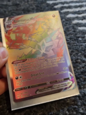 Pokémonkort Eternatus VMAX Full Art - Säljer ett Pokémonkort av modellen Eternatus VMAX Full Art med holografisk yta. Kortet har färgglada geometriska mönster i regnbågsfärger och text på engelska. Perfekt för samlare eller Pokémon-fans som vill utöka sin samling.
