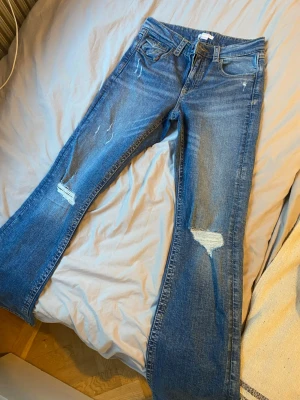 Blå bootcut jeans med slitningar - Snygga blå bootcut jeans med slitna detaljer på benen och klassiska fem fickor. Jeansen har låg midja och utställda ben, samt dekorativa bakfickor med knapp. Perfekta för en avslappnad och trendig look.
