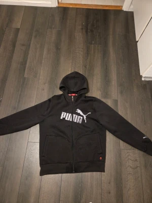 Svart hoodie från Puma med dragkedja - Svart hoodie från Puma med dragkedja framtill och stor vit logga på bröstet. Tröjan har huva, två fickor fram och ribbade muddar vid ärmslut och nederkant. Perfekt för en sportig och avslappnad stil.