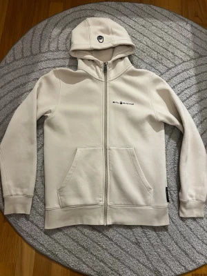 Vit zip-hoodie från Sail Racing - Snygg vit hoodie med dragkedja från Sail Racing. Tröjan har huva med broderad logga, två stora fickor fram och diskret Sail Racing-tryck på bröstet. Perfekt för en clean och stilren look. Mjuk bomullskänsla och ribbade muddar.