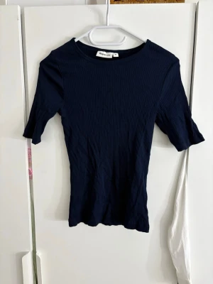 Mörkblå ribbad topp från Kappahl - En mörkblå ribbad topp från Kappahl med rund halsringning och halvlånga ärmar. Toppen är figurnära och har en stretchig känsla, perfekt att styla med jeans eller kjol. Materialet är mjukt och bekvämt, och den ribbade strukturen ger en snygg detalj.