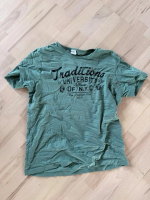 Grön t-shirt med tryck NYC - Grön t-shirt från Traditions med svart tryck på bröstet där det står 'Traditions University of N.Y.C'. Klassisk rund hals och korta ärmar. Mjuk bomullskänsla och avslappnad passform, perfekt för en chill vardagslook.