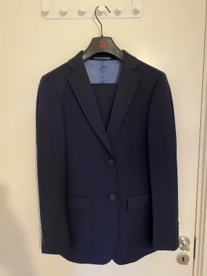 Dressman Kostym - Dressmans bäst säljande kostym och byxor i marin blå färg. Har bara andvänts 2 gånger och är i nyskick utan några fläckar eller skador. Kavajen är i storlek 44 och byxorna är 30-32. Kavaj eller byxa går att köpas separat ifall det önskas, bara skicka ett meddelande så löser jag det utan problem. 