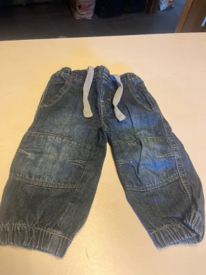 Mörkblå jeans med resår och snörning - Säljer ett par mörkblå jeans med resår i midjan och benslut samt grå snörning framtill. Jeansen har flera fickor och förstärkta sömmar på benen för en cool look. Perfekta för en avslappnad och bekväm stil.