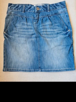 Low waist jeanskjol i stretch – Y2K-stil från jsfn denim (strl 36) - Snygg jeanskjol i klassisk blå denim med bekväm stretch från jsfn (Ellos). Modellen är low waist (låg midja) med lätta veck upptill, gylf med dragkedja, dekorativ knapp och fungerande fickor fram och bak. Mycket gott skick, perfekt för en trendig Y2K-look! Mått (mätt platt): • Midjebredd: ca 39 cm (sitter på höften då det är low waist) • Midjeomkrets: ca 78 cm • Längd: 44 cm från midjan och ner Material: 97% Bomull, 3% Elastan.