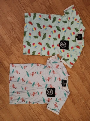 Två t-shirts med coola tryck herr stl XS - Två coola t-shirts från Blount&Pool. Ena t-shirten är ljusgrön med mönster av glassar, vattenmeloner och krokodiler. Den andra är ljusblå med tryck av flamingos och surfbrädor. Båda har korta ärmar och är i ekologisk bomull. Perfekta för sommaren! Båda är i herr stl XS. Bra skick.