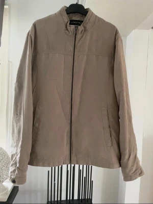 Beige jacka från Zara Man - Stilren beige jacka från Zara Man med rak passform och ståkrage. Jackan har dragkedja framtill, två sidofickor och knappdetaljer vid ärmsluten. Tillverkad i ett mjukt bomullsliknande material som passar perfekt till vardagslooken.