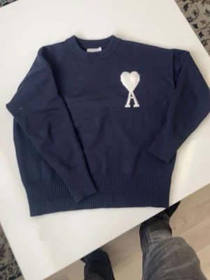  Ami Paris tröja - Mörkblå stickad tröja från Ami Paris med rund halsringning och ribbade muddar. På bröstet finns ett vitt hjärta med bokstaven A broderat. Perfekt för dig som gillar stilrena och trendiga plagg.