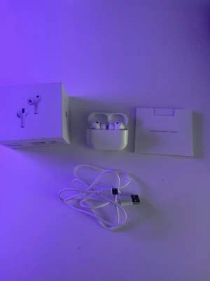 Airpods  - Trådlösa vita in-ear hörlurar från Apple, modell AirPods Pro, med tillhörande laddningsetui och USB-laddningskabel. Snygg och stilren design i blankt plastmaterial. Levereras med originalförpackning och dokumentation.