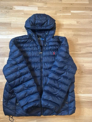 Mörkblå vårjacka Polo Ralph Lauren - Snygg mörkblå pufferjacka från Polo Ralph Lauren med röd broderad logga på bröstet. Jackan har huva, dragkedja framtill och två fickor med dragkedja. Perfekt för kalla dagar och riktigt skön att bära. Skick 10/10 använd en gång