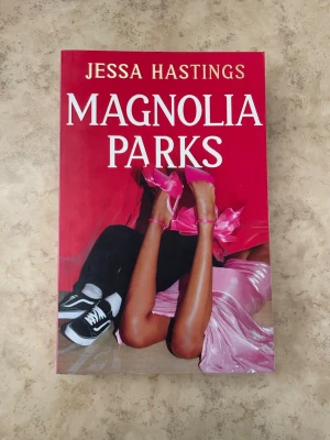 Magnolia Parks av Jessa Hastings - Magnolia Parks är en skönlitterär bok med ett färgstarkt omslag i rosa och rött. Omslaget visar ben i rosa klackar och sneakers mot en bakgrund av rosa tyg. Perfekt för dig som gillar ungdomsromaner och drama.
