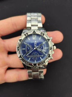 Blå och silverfärgad klocka Geneva - Stilren klocka från Geneva med blå urtavla och silverfärgat armband i metall. Klockan har tydliga siffror, flera dekorativa urtavlor och en robust boett med markerade siffror runt ringen. Perfekt accessoar för dig som gillar sportig och modern stil. Ända att klaga på är att minutvisaren är lite lös när man vrider på klockan.