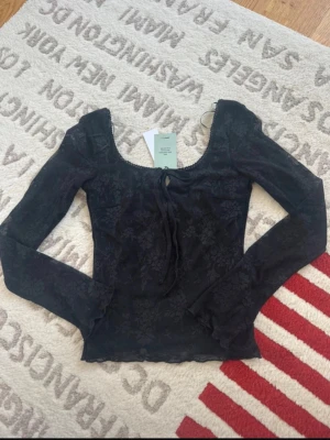 Svart blommig mesh topp från Gina Tricot - Trendig svart mesh topp från Gina Tricot med blommigt mönster och långa ärmar. Toppen har en djup rundringning och knyt framtill för extra detalj. Perfekt att styla med jeans eller kjol för en cool look.