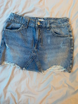 Blå jeanskjol med fransig kant - Snygg blå jeanskjol med fransig och rå kant nertill. Kjolen har klassiska fickor fram och bak, bälteshällor och knappstängning. Perfekt för en avslappnad och trendig look.